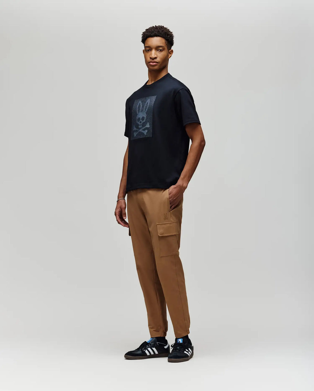 A man wearing the Psycho Bunny MENS REECE OVERSIZED TEE (MT0200082) in black, tan cargo pants, and black Adidas sneakers stands against a plain light gray background, looking toward the camera with a neutral expression.