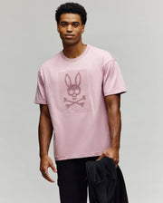 A man in a Psycho Bunny MENS REECE OVERSIZED TEE - MT0200082, a relaxed fit light pink Pima cotton shirt with bunny and crossbones graphic, stands against a plain background holding a black jacket and smiling slightly at the camera.