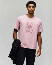 A man faces forward holding a black jacket, wearing the Psycho Bunny MENS REECE OVERSIZED TEE (MT0200082) in light pink Pima cotton with a skull and crossbones bunny logo, set against a plain light gray background.