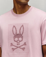 A person wears the Psycho Bunny MENS REECE OVERSIZED TEE (MT0200082), a light pink Pima cotton shirt with an embossed bunny skull and crossbones subtly blended into the fabric; their face is partially hidden, highlighting the tee’s relaxed, premium style.