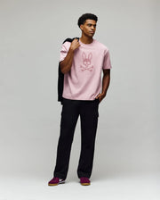 A man stands against a plain background in the Psycho Bunny MENS REECE OVERSIZED TEE - MT0200082, a relaxed fit light pink Pima cotton shirt with bunny and crossbones graphic, paired with black cargo pants, maroon sneakers, and a black jacket over his shoulder.