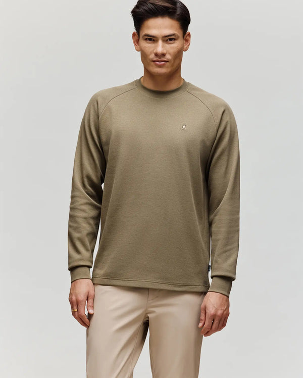 A man stands against a plain background wearing the Psycho Bunny MENS EVANS WAFFLE (MT0200091) shirt in khaki, featuring a signature embroidered Bunny logo on the left chest, paired with light beige pants and a neutral expression.