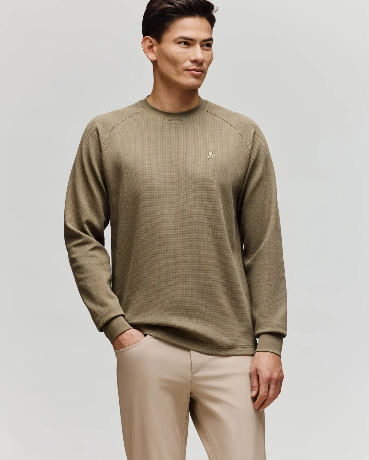 A man stands against a plain background wearing Psycho Bunny’s MENS EVANS WAFFLE - MT0200091 light olive green waffle knit long-sleeve sweater and beige pants, with one hand in his pocket and a relaxed expression.