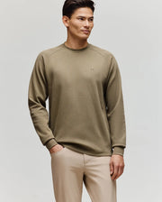 A man stands against a plain background wearing Psycho Bunny’s MENS EVANS WAFFLE - MT0200091 light olive green waffle knit long-sleeve sweater and beige pants, with one hand in his pocket and a relaxed expression.