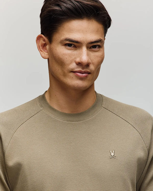 A man with short dark hair wears the Psycho Bunny MENS EVANS WAFFLE - MT0200091 sweater, featuring a light olive waffle knit and embroidered Bunny logo, standing against a plain light background and looking slightly to the side with a subtle smile.