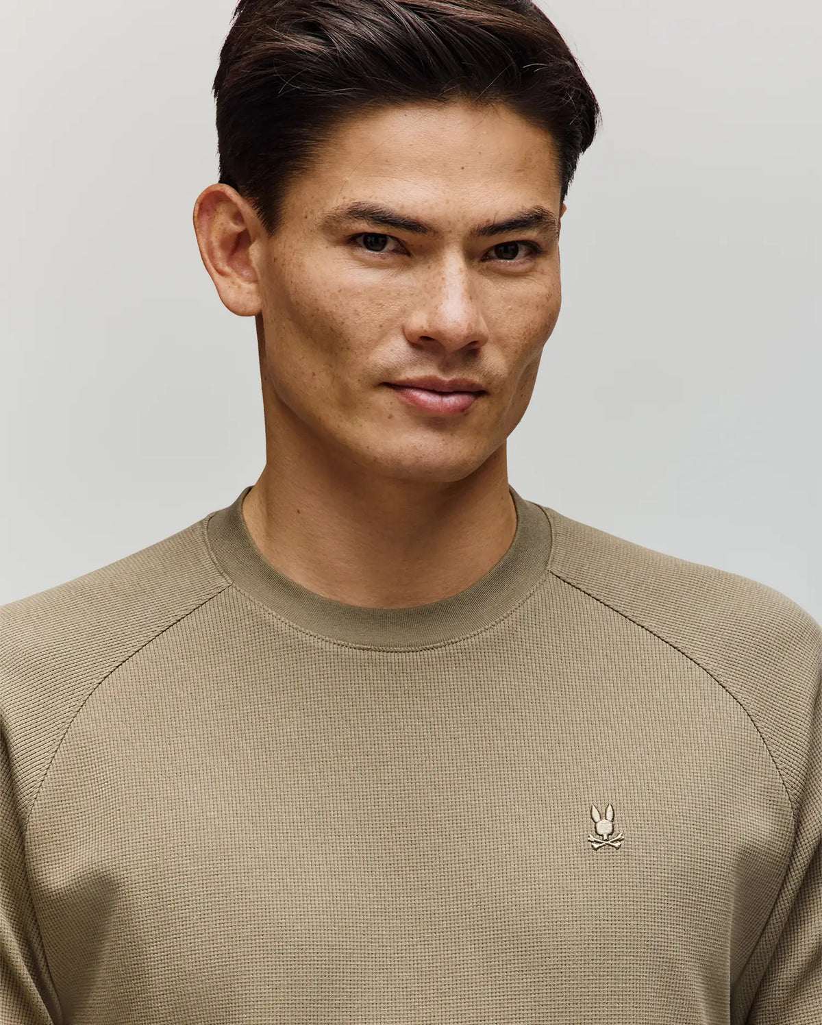 A man with short dark hair wears the Psycho Bunny MENS EVANS WAFFLE - MT0200091 sweater, featuring a light olive waffle knit and embroidered Bunny logo, standing against a plain light background and looking slightly to the side with a subtle smile.