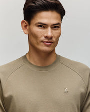 A man with short dark hair wears the Psycho Bunny MENS EVANS WAFFLE - MT0200091 sweater, featuring a light olive waffle knit and embroidered Bunny logo, standing against a plain light background and looking slightly to the side with a subtle smile.