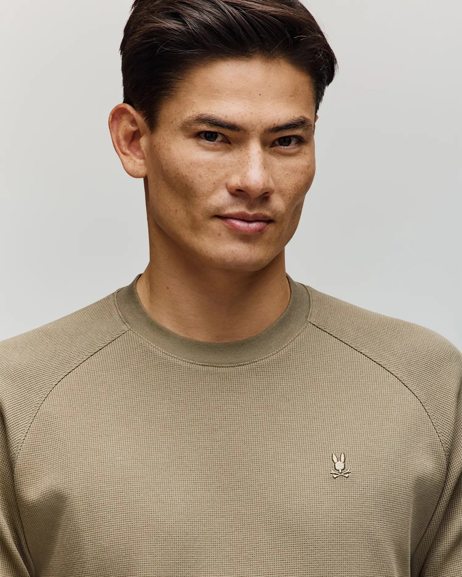 A man with short dark hair wears the Psycho Bunny MENS EVANS WAFFLE - MT0200091 sweater, featuring a light olive waffle knit and embroidered Bunny logo, standing against a plain light background and looking slightly to the side with a subtle smile.