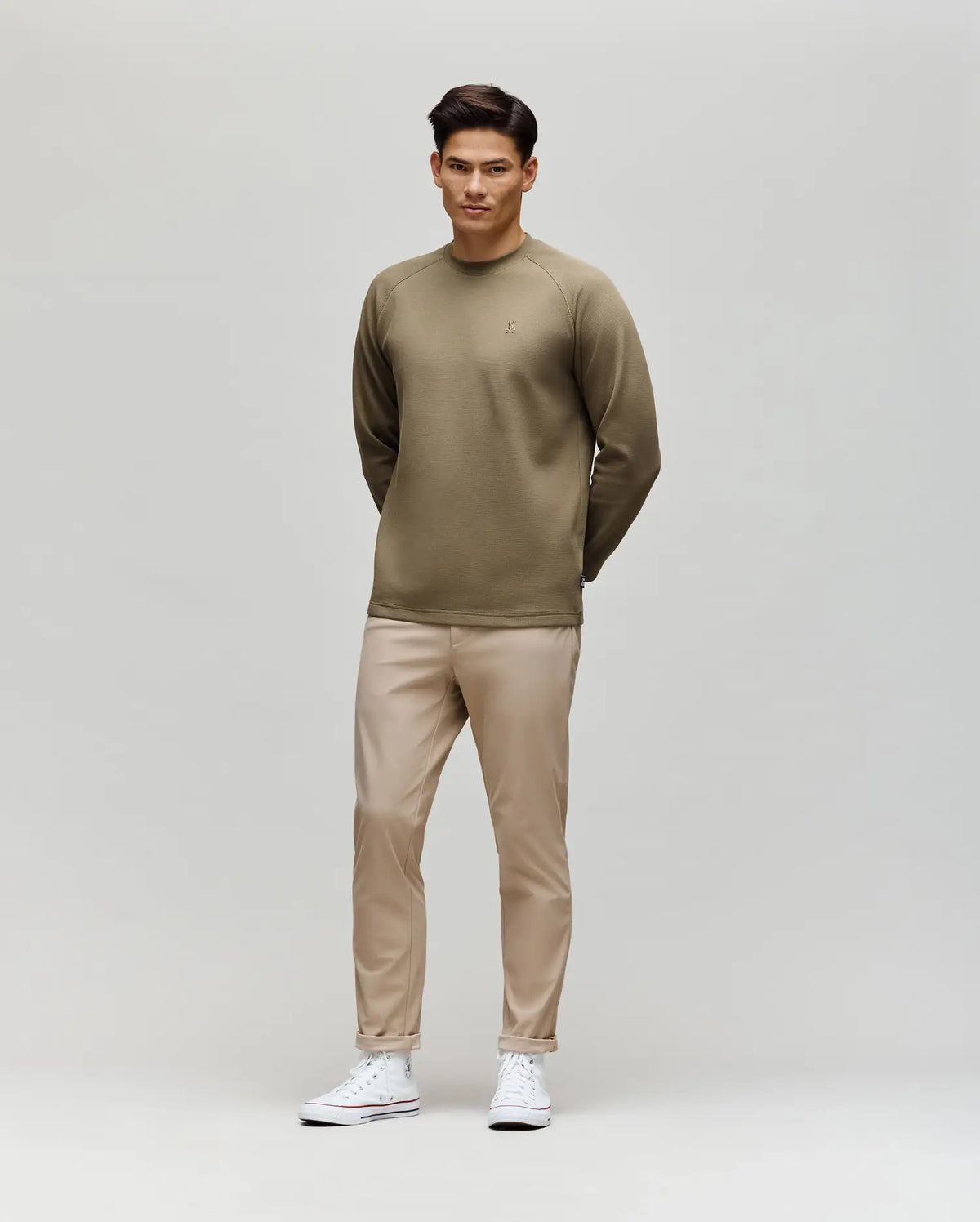 A man stands against a light background wearing the Psycho Bunny MENS EVANS WAFFLE - MT0200091 khaki shirt with an embroidered Bunny, beige pants, and white sneakers. His hands are behind his back and he looks forward neutrally.