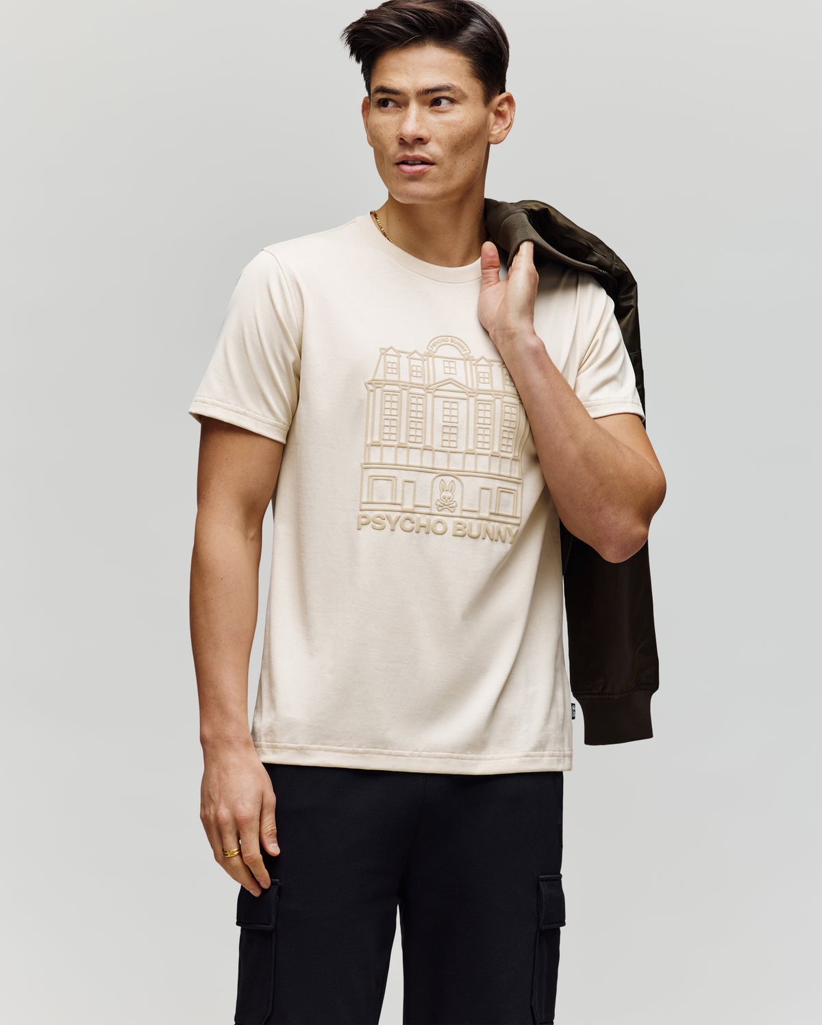 A man wears the Psycho Bunny MENS REDFORD GRAPHIC TEE (MT0200092), a light beige Pima cotton shirt with an outlined building and "Psycho Bunny" text, paired with black pants and holding a dark jacket against a plain background.
