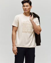 A man wears the Psycho Bunny MENS REDFORD GRAPHIC TEE (MT0200092), a light beige Pima cotton shirt with an outlined building and "Psycho Bunny" text, paired with black pants and holding a dark jacket against a plain background.