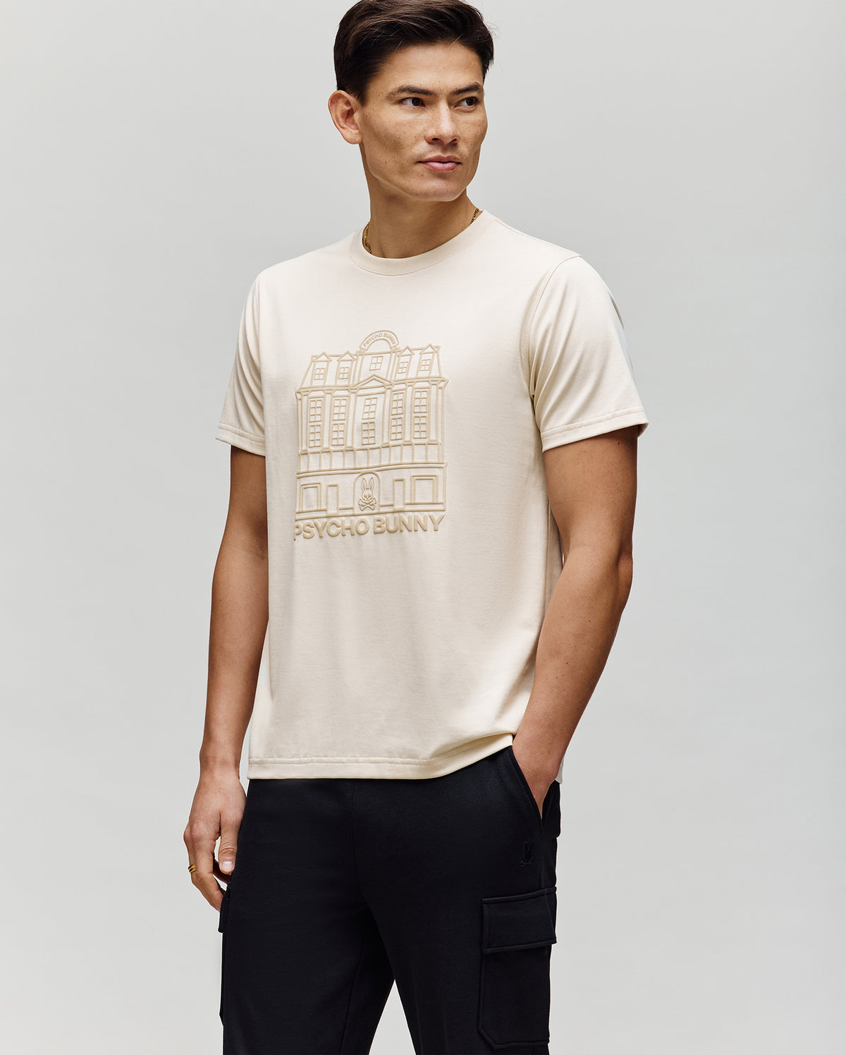 A man wears the Psycho Bunny MENS REDFORD GRAPHIC TEE - MT0200092, a cream-colored Pima cotton shirt featuring a building line drawing and "PSYCHO BUNNY" on the front, paired with black pants against a plain light background.
