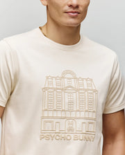 A person wears the Psycho Bunny MENS REDFORD GRAPHIC TEE (MT0200092), a cream Pima cotton shirt featuring an embossed building design, "PSYCHO BUNNY" text, and a small bunny logo; their face is partially out of frame.