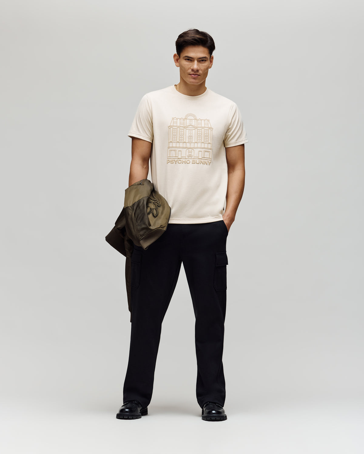 A man stands against a plain background, wearing a light-colored Psycho Bunny MENS REDFORD GRAPHIC TEE - MT0200092, black cargo pants, and black shoes. He holds an olive green jacket in his left hand and gazes slightly left with a neutral expression.