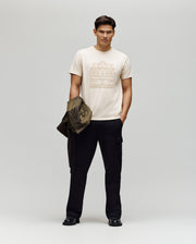 A man stands against a plain background, wearing a light-colored Psycho Bunny MENS REDFORD GRAPHIC TEE - MT0200092, black cargo pants, and black shoes. He holds an olive green jacket in his left hand and gazes slightly left with a neutral expression.