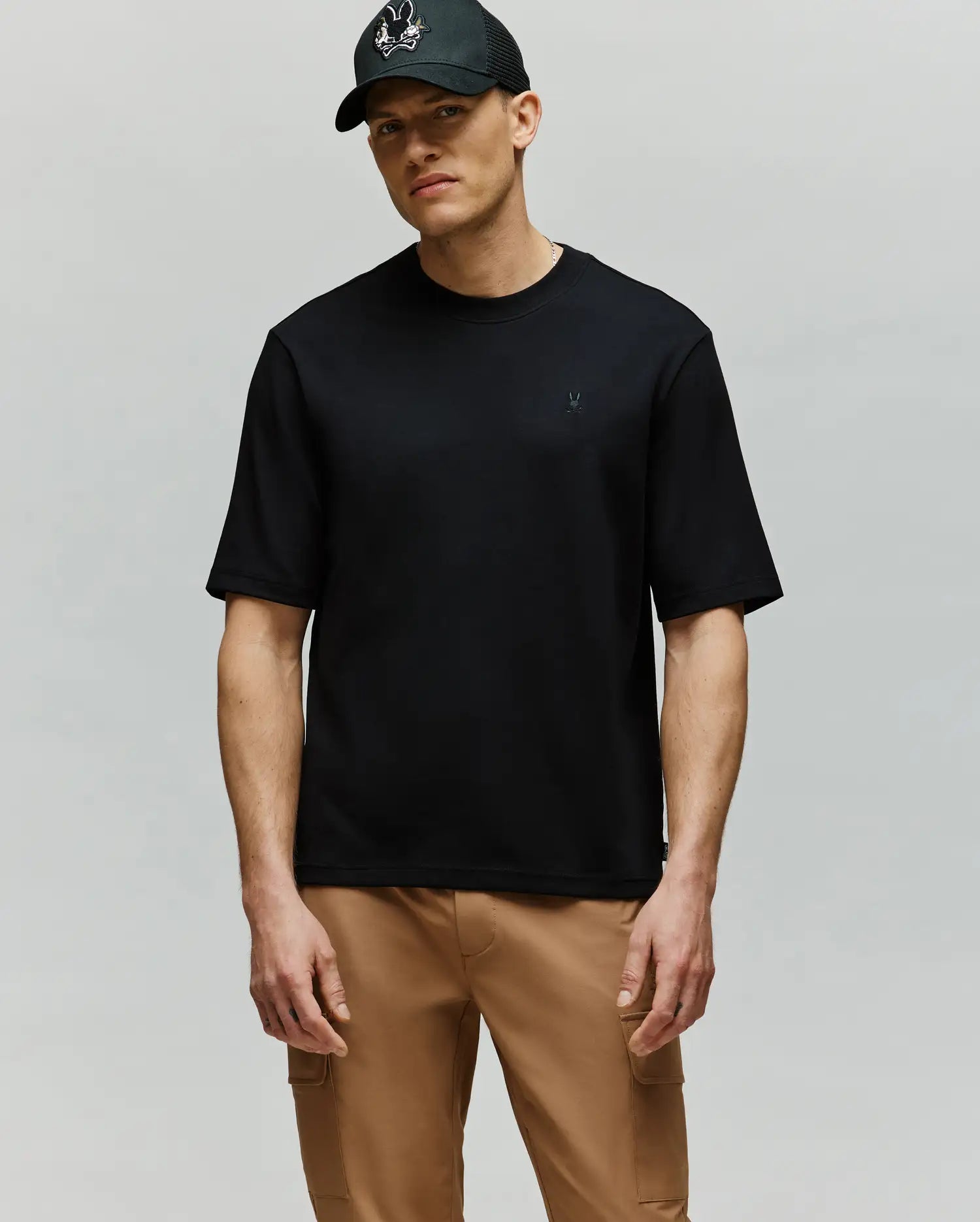 A man wearing a black cap, tan cargo pants, and the Psycho Bunny MENS ADAM INTERLOCK TEE (MT0200093) with an embroidered Bunny logo stands against a light grey background, looking forward with a neutral expression.