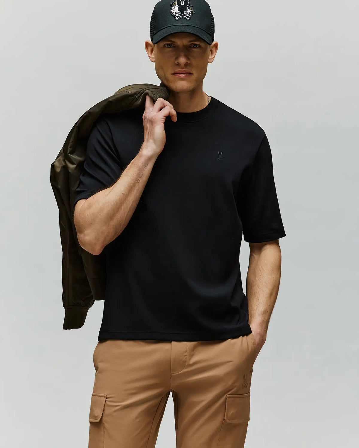A man in a black cap, tan cargo pants, and the Psycho Bunny MENS ADAM INTERLOCK TEE (MT0200093) with an embroidered Bunny logo holds a jacket over his shoulder, standing against a plain light gray background.