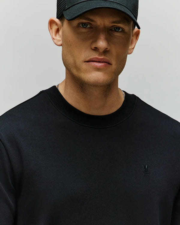 A man wears the Psycho Bunny MENS ADAM INTERLOCK TEE (MT0200093) featuring a small embroidered Bunny logo on the chest, and a black baseball cap, looking slightly serious against a plain background.