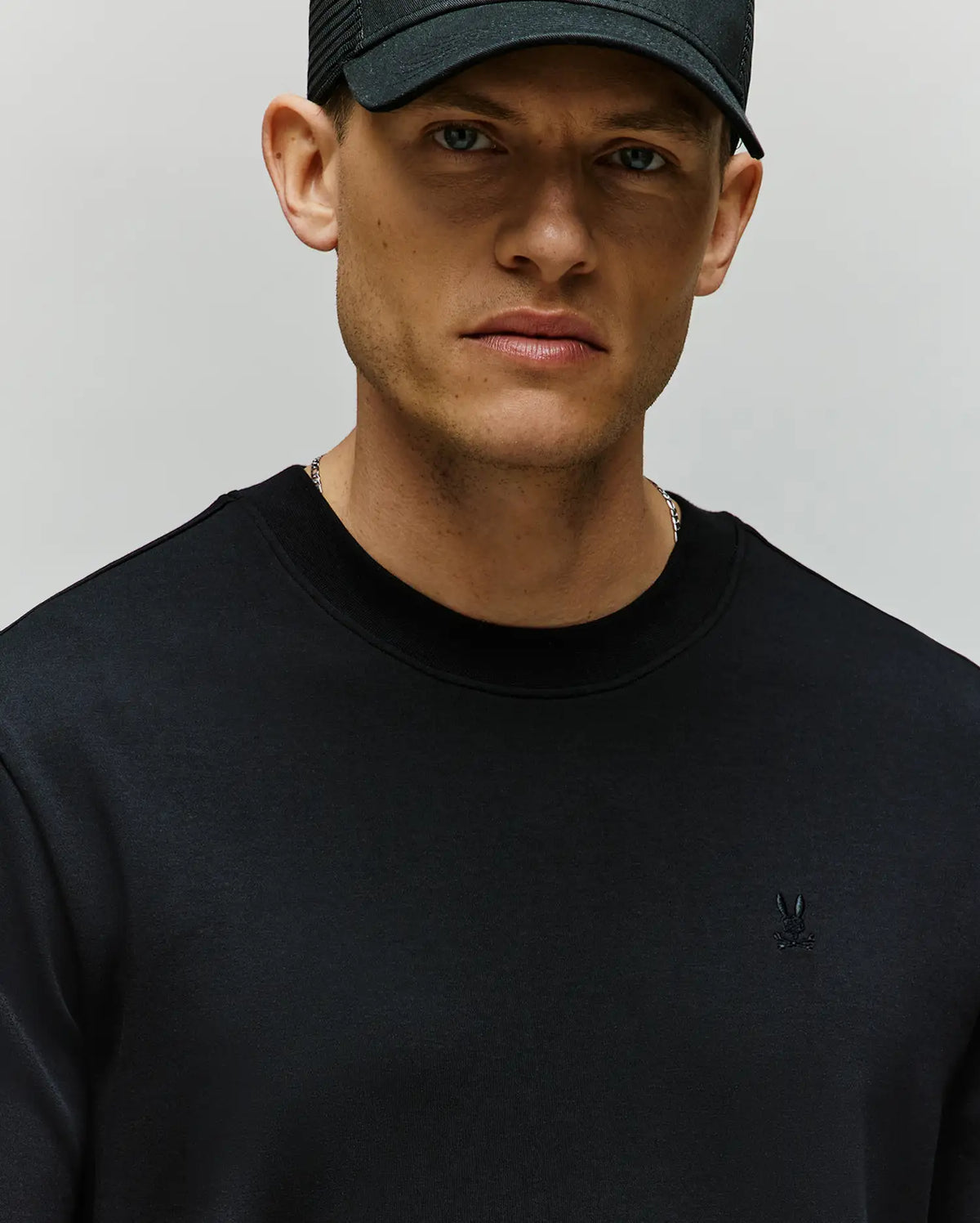 A man wears the Psycho Bunny MENS ADAM INTERLOCK TEE (MT0200093) featuring a small embroidered Bunny logo on the chest, and a black baseball cap, looking slightly serious against a plain background.