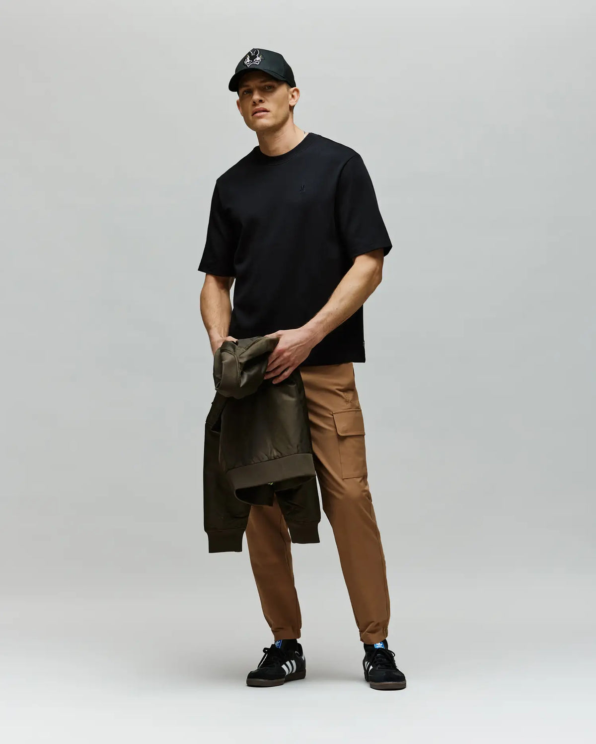 A man wears the Psycho Bunny MENS ADAM INTERLOCK TEE (MT0200093) in black, tan joggers, and black sneakers, holding a green jacket in his left hand against a plain gray background.