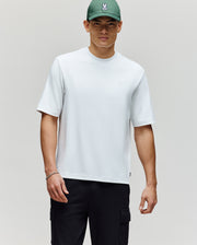A man in a Psycho Bunny MENS ADAM INTERLOCK TEE (MT0200093), black cargo pants, and a green cap with an embroidered Bunny logo stands against a light gray background.