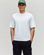 A man stands against a neutral background with his hands in his pockets, wearing the Psycho Bunny MENS ADAM INTERLOCK TEE (MT0200093), black cargo pants, and a green cap featuring an embroidered Bunny logo.