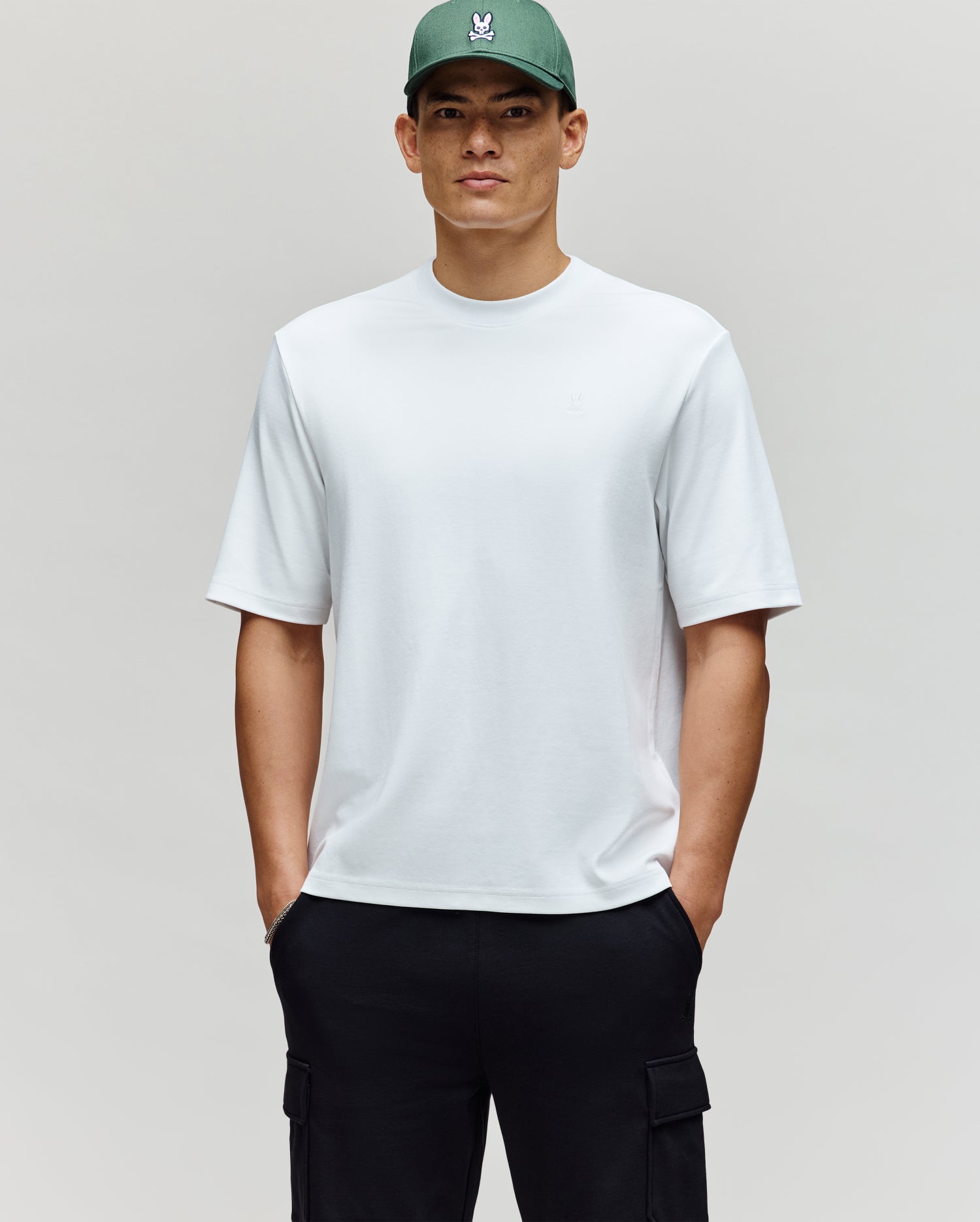 A man stands against a neutral background with his hands in his pockets, wearing the Psycho Bunny MENS ADAM INTERLOCK TEE (MT0200093), black cargo pants, and a green cap featuring an embroidered Bunny logo.