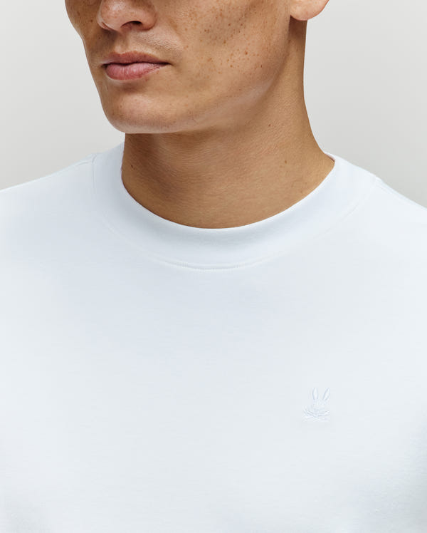 A close-up of a person wearing the Psycho Bunny MENS ADAM INTERLOCK TEE - MT0200093, a plain white men’s knit t-shirt with a subtle white embroidered Bunny logo on the chest; only the lower half of the face and upper torso are visible.