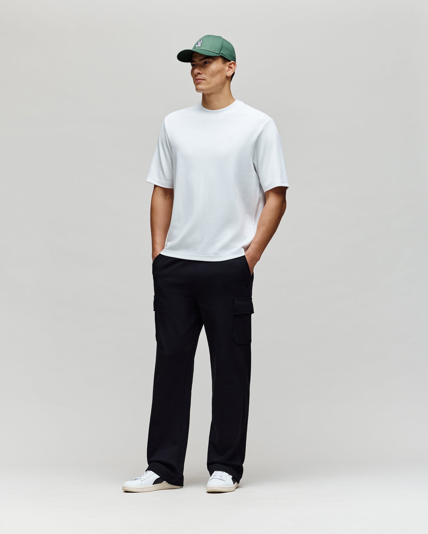 A man stands against a plain light background wearing the Psycho Bunny MENS ADAM INTERLOCK TEE (MT0200093)—a white knit t-shirt with an embroidered Bunny logo—paired with black cargo pants, white sneakers, and a green baseball cap, hands in pockets.