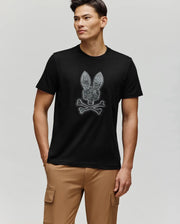 A man wearing the Psycho Bunny MENS ROWEN BUNNY GRAPHIC TEE (MT0200101)—a black Pima cotton shirt with a bunny skull and crossbones—stands with one hand in his tan cargo pants pocket against a plain light gray background.