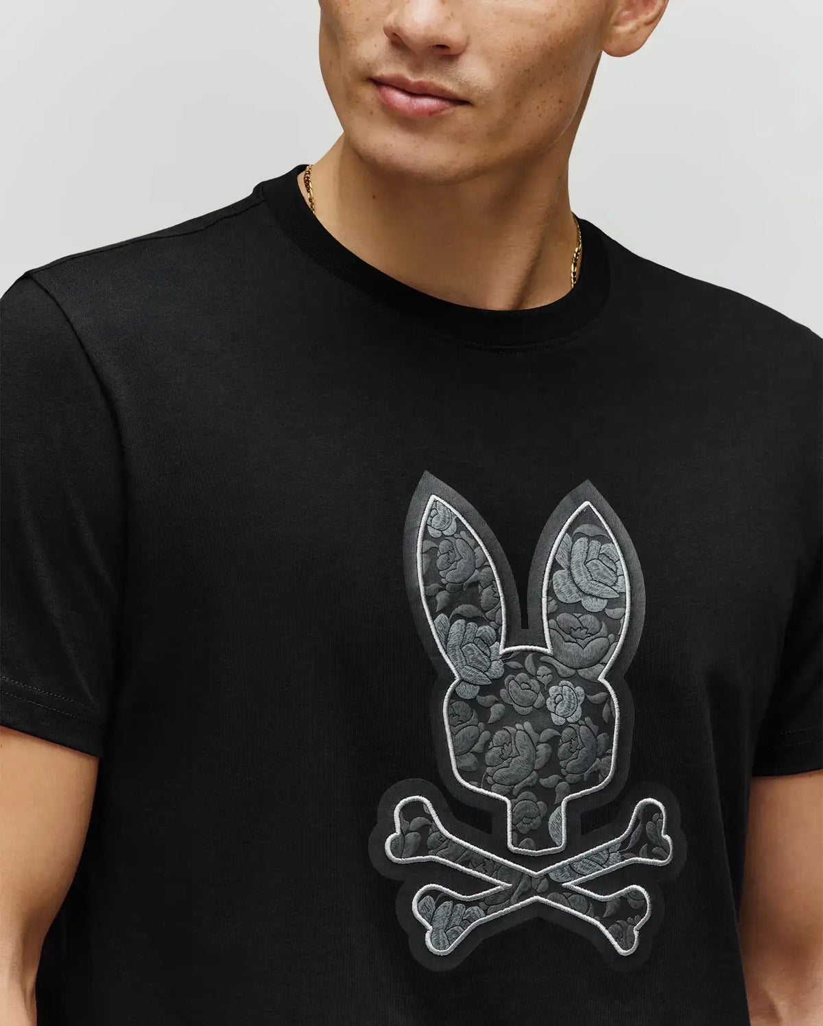 A person is seen from the lower face to upper torso wearing the Psycho Bunny MENS ROWEN BUNNY GRAPHIC TEE (MT0200101), a black Pima cotton shirt with a bold bunny graphic, crossbones, and floral pattern.