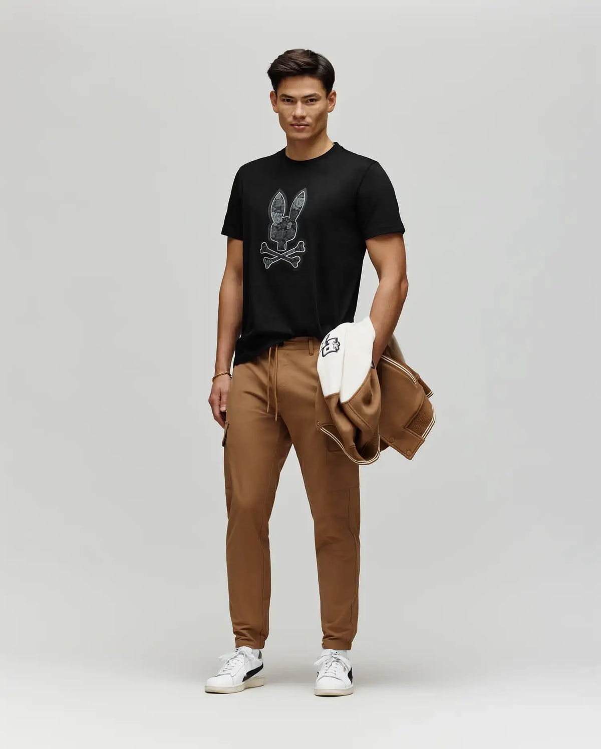 A man faces the camera against a plain background, wearing the Psycho Bunny MENS ROWEN BUNNY GRAPHIC TEE - MT0200101 in black Pima cotton with a bunny graphic, brown jogger pants, and white sneakers. He holds a tan jacket and cap in his left hand.