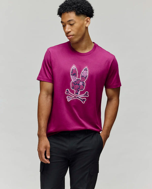 A man wears the Psycho Bunny MENS ROWEN BUNNY GRAPHIC TEE - MT0200101, featuring an embroidered Bunny with floral accents on soft Pima cotton, paired with black pants against a plain light gray background.