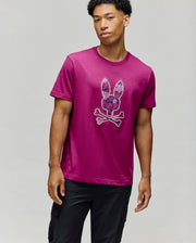A young man with short curly hair wears the Psycho Bunny MENS ROWEN BUNNY GRAPHIC TEE - MT0200101 in magenta, featuring an embroidered Bunny graphic, styled with black cargo pants against a plain light gray background.