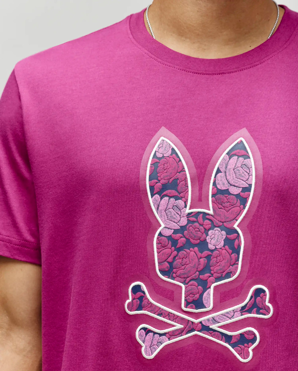 A person wears the Psycho Bunny MENS ROWEN BUNNY GRAPHIC TEE (MT0200101) in magenta, made from soft Pima cotton with an embroidered Bunny graphic featuring a pink and purple rose pattern, paired with a visible silver chain necklace.