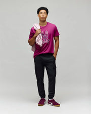 A man poses against a plain background in the Psycho Bunny MENS ROWEN BUNNY GRAPHIC TEE (MT0200101) in magenta, made from soft Pima cotton with an embroidered Bunny graphic, black cargo pants, magenta sneakers, and a light pink jacket over his shoulder.