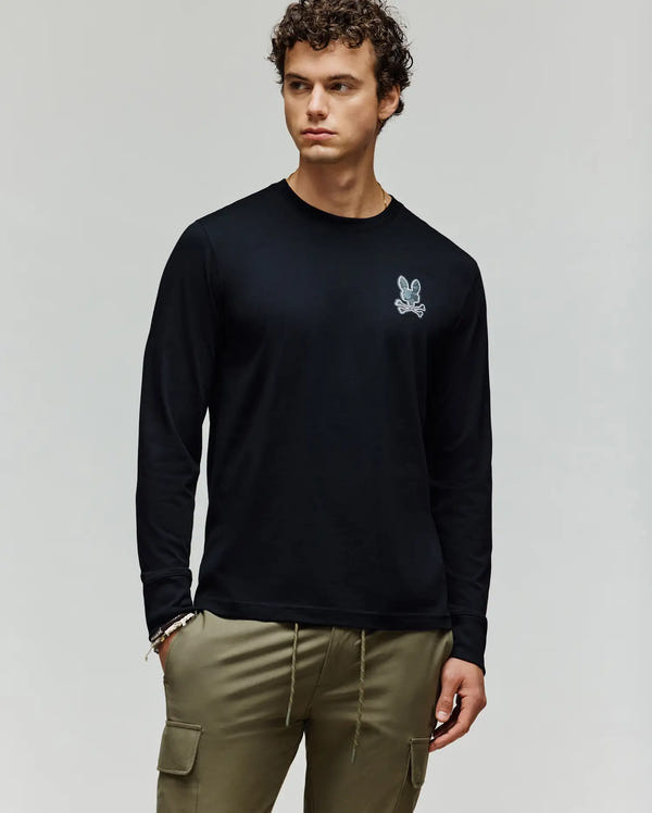 A man with curly hair wears a black Psycho Bunny MENS ROWEN GRAPHIC TEE - MT0200103 featuring a small white chest graphic, paired with olive green cargo pants, standing against a plain light background.