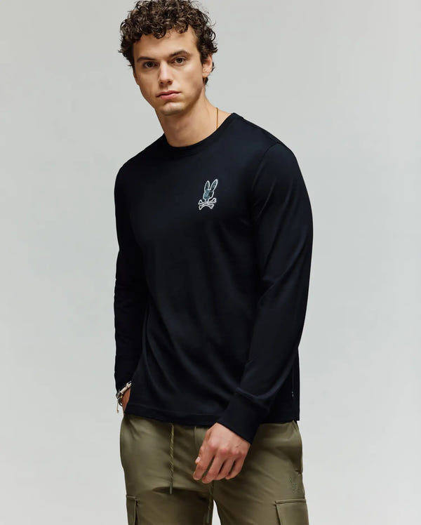 A man with curly hair wears Psycho Bunny’s MENS ROWEN GRAPHIC TEE - MT0200103, a black long sleeve Pima cotton shirt with a small white bunny logo on the chest, paired with olive green pants against a light gray background.