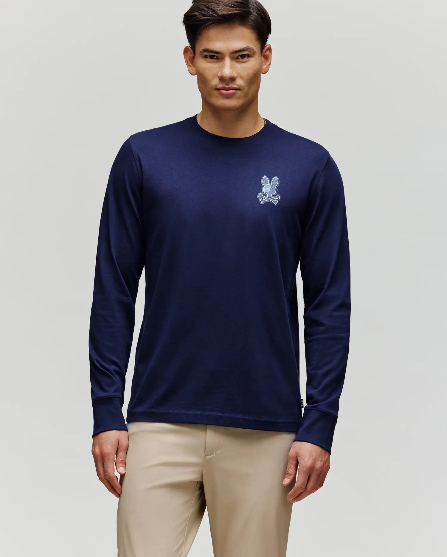 A person wears a navy MENS ROWEN GRAPHIC TEE (MT0200103) by Psycho Bunny, featuring a small white bunny logo on the chest, paired with light beige pants, standing against a plain light background.