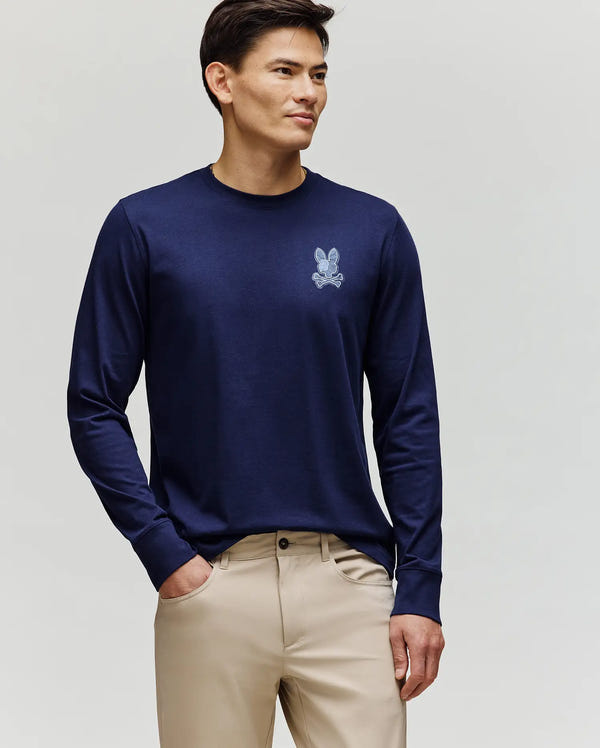 A man stands against a light background wearing the Psycho Bunny MENS ROWEN GRAPHIC TEE (MT0200103) in navy blue, featuring a small bunny logo on the chest, paired with light beige pants.