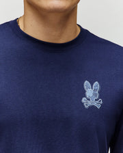 A person wearing a Psycho Bunny MENS ROWEN GRAPHIC TEE (MT0200103) in navy, featuring a light blue bunny skull and crossbones logo on the left chest. Only their lower face, neck, and upper torso are visible.