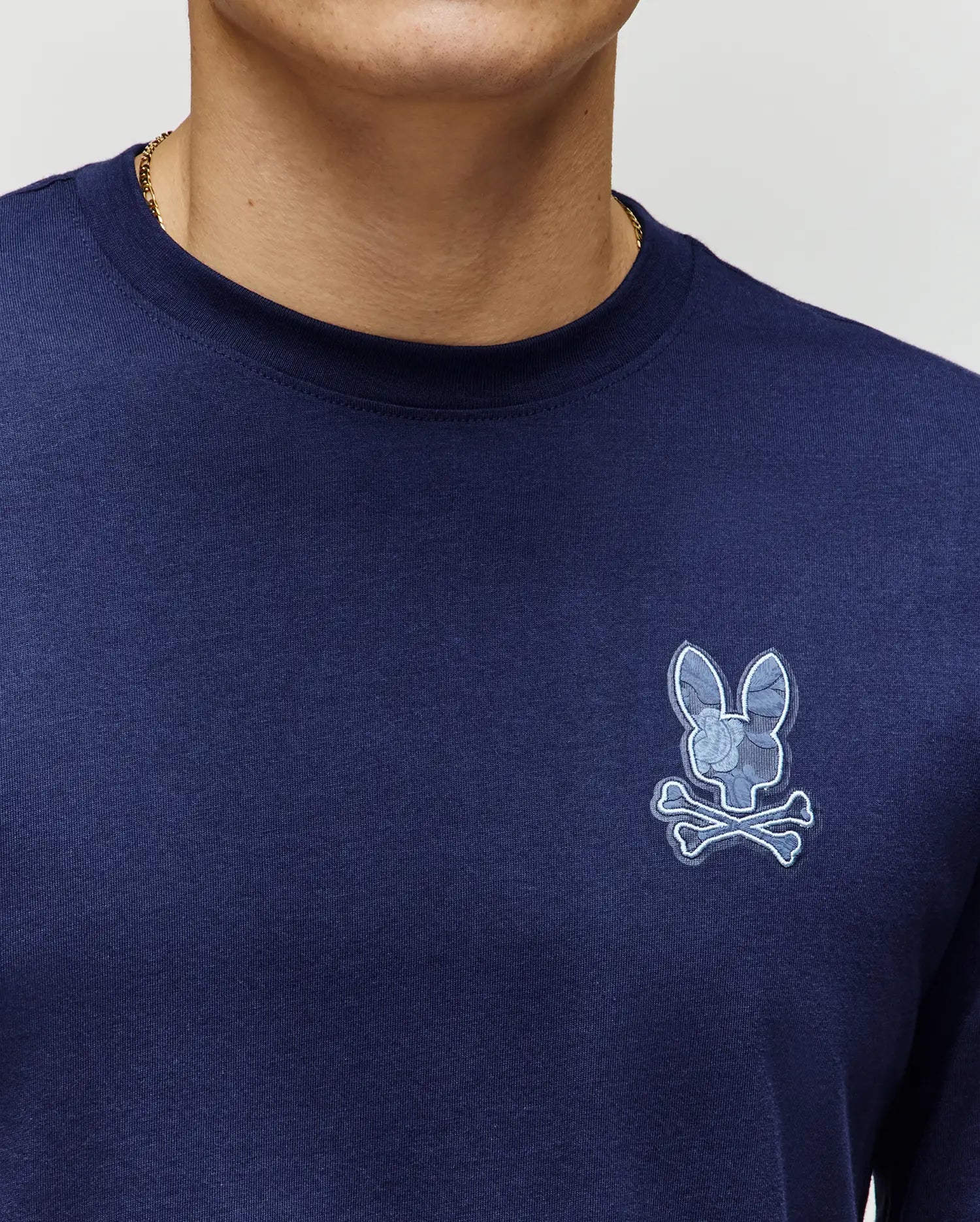 A person wearing a Psycho Bunny MENS ROWEN GRAPHIC TEE (MT0200103) in navy, featuring a light blue bunny skull and crossbones logo on the left chest. Only their lower face, neck, and upper torso are visible.