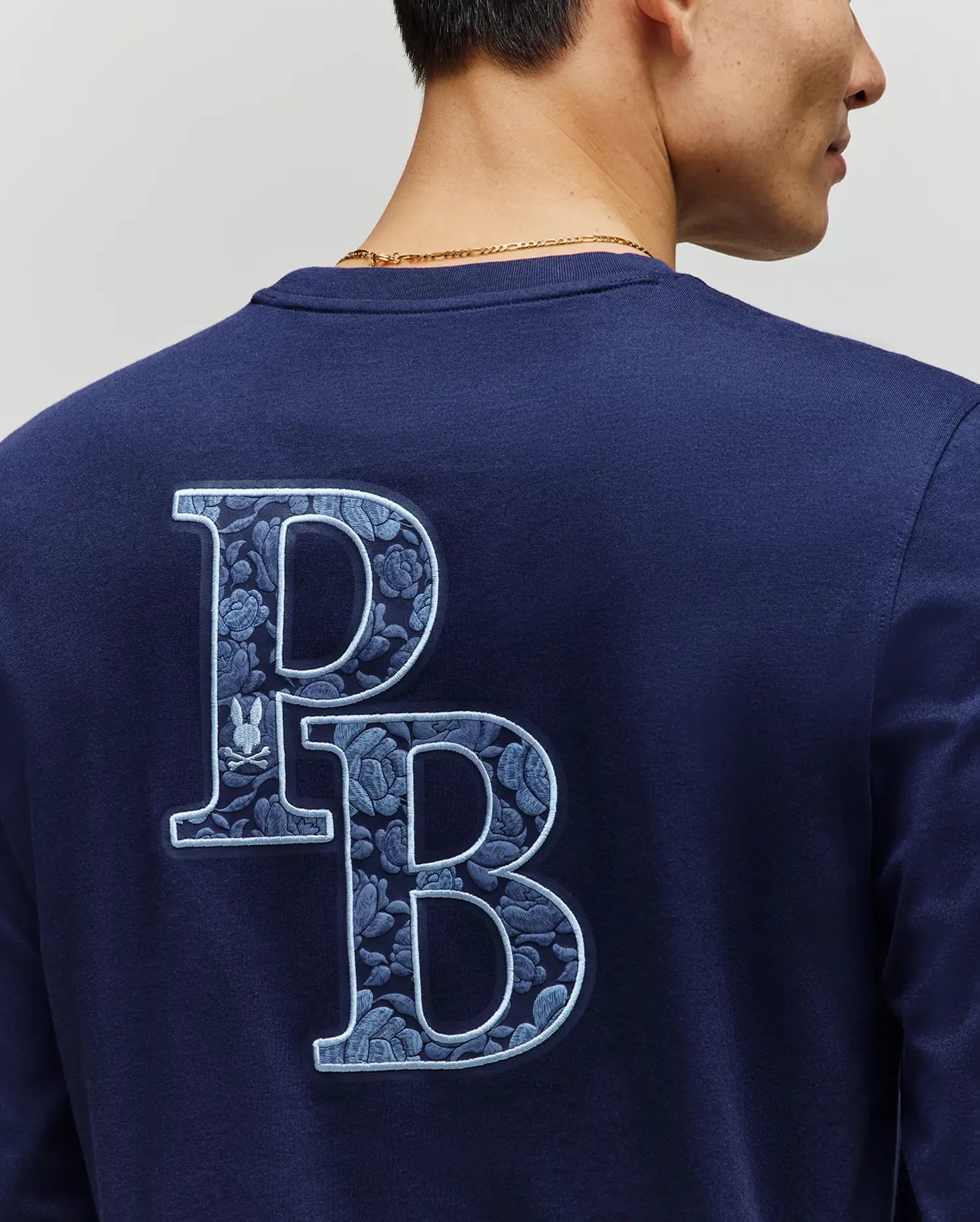 A person faces away, showing only neck, shoulder, and part of their face while wearing the Psycho Bunny MENS ROWEN GRAPHIC TEE (MT0200103) in navy blue with large light blue floral "PB" letters on the back.