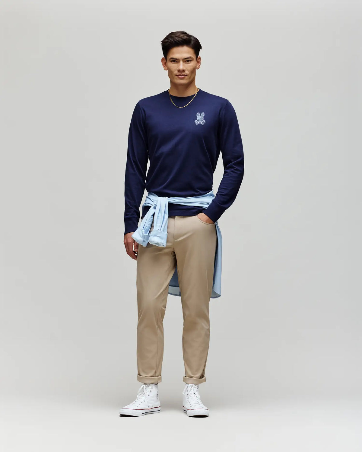 A man stands against a plain background wearing a navy Psycho Bunny MENS ROWEN GRAPHIC TEE (MT0200103) with a small logo, beige pants, white high-top sneakers, a light blue shirt tied at his waist, and a gold chain necklace.