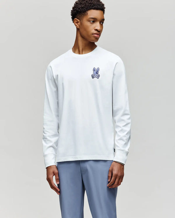 A young man in a Psycho Bunny MENS ROWEN GRAPHIC TEE - MT0200103, featuring a white Pima cotton build with a small blue cartoon rabbit on the chest, and light blue pants, stands against a plain gray background looking to the side.