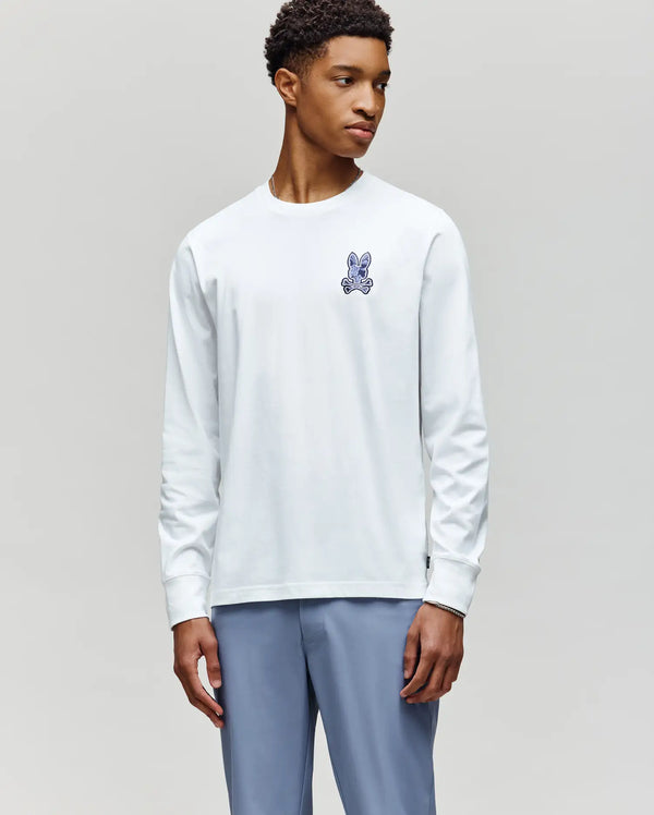 A young man in a Psycho Bunny MENS ROWEN GRAPHIC TEE - MT0200103, featuring a white Pima cotton build with a small blue cartoon rabbit on the chest, and light blue pants, stands against a plain gray background looking to the side.