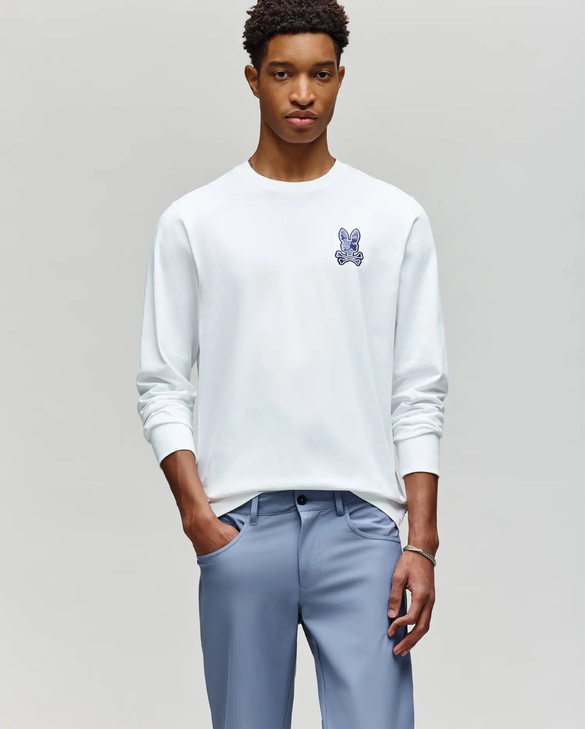 A young man stands against a plain background, wearing the Psycho Bunny MENS ROWEN GRAPHIC TEE (MT0200103) in white with a small blue bunny logo on the chest and light blue pants, hands in his pockets as he looks at the camera.