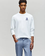 A young man stands against a plain background, wearing the Psycho Bunny MENS ROWEN GRAPHIC TEE (MT0200103) in white with a small blue bunny logo on the chest and light blue pants, hands in his pockets as he looks at the camera.