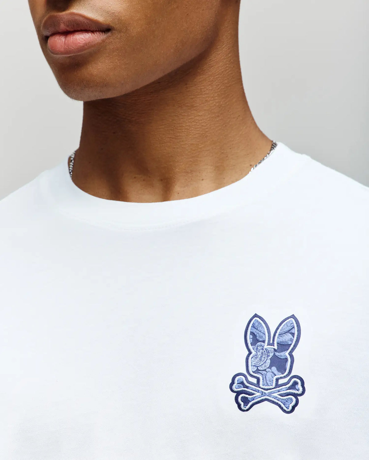 A person wears the Psycho Bunny MENS ROWEN GRAPHIC TEE - MT0200103, a white long sleeve shirt with a blue patch and the iconic bunny face and crossbones graphic on the left chest; only their lower face and upper torso are shown.