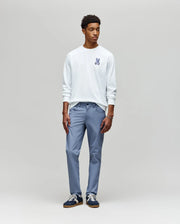 A young man stands against a plain light gray background, wearing the Psycho Bunny MENS ROWEN GRAPHIC TEE (MT0200103) in white, paired with light blue pants and black sneakers with white soles, looking slightly to his left.